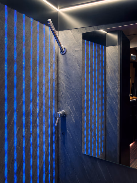 Dusche im VANYX mit dunkler Steinwand, vertikalen blauen LED-Leisten, Handbrause und Spiegel